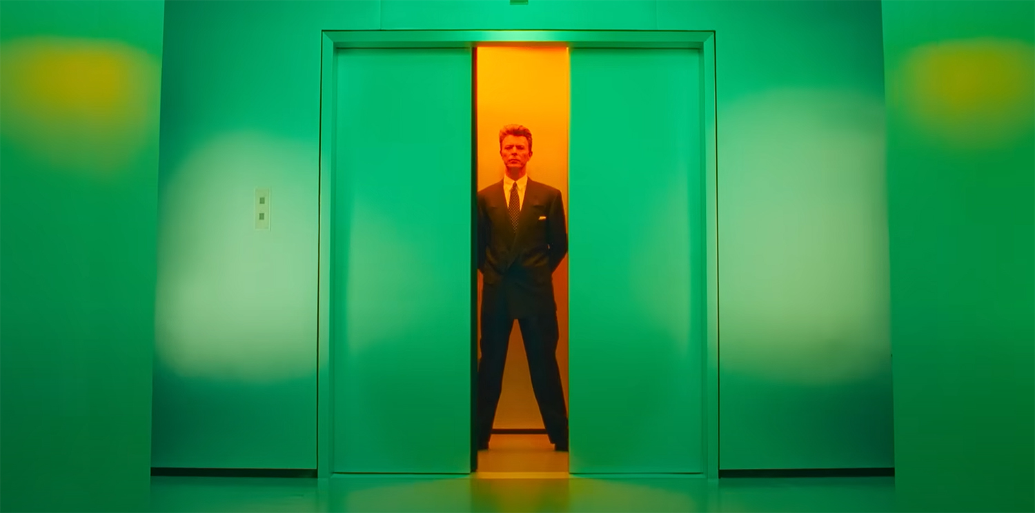 Bowie stands in an orange lit elevator as the green doors close.
