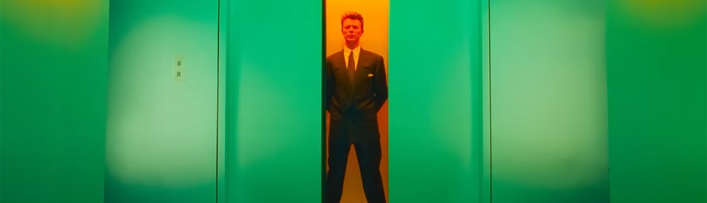 Bowie stands in an orange lit elevator as the green doors close.
