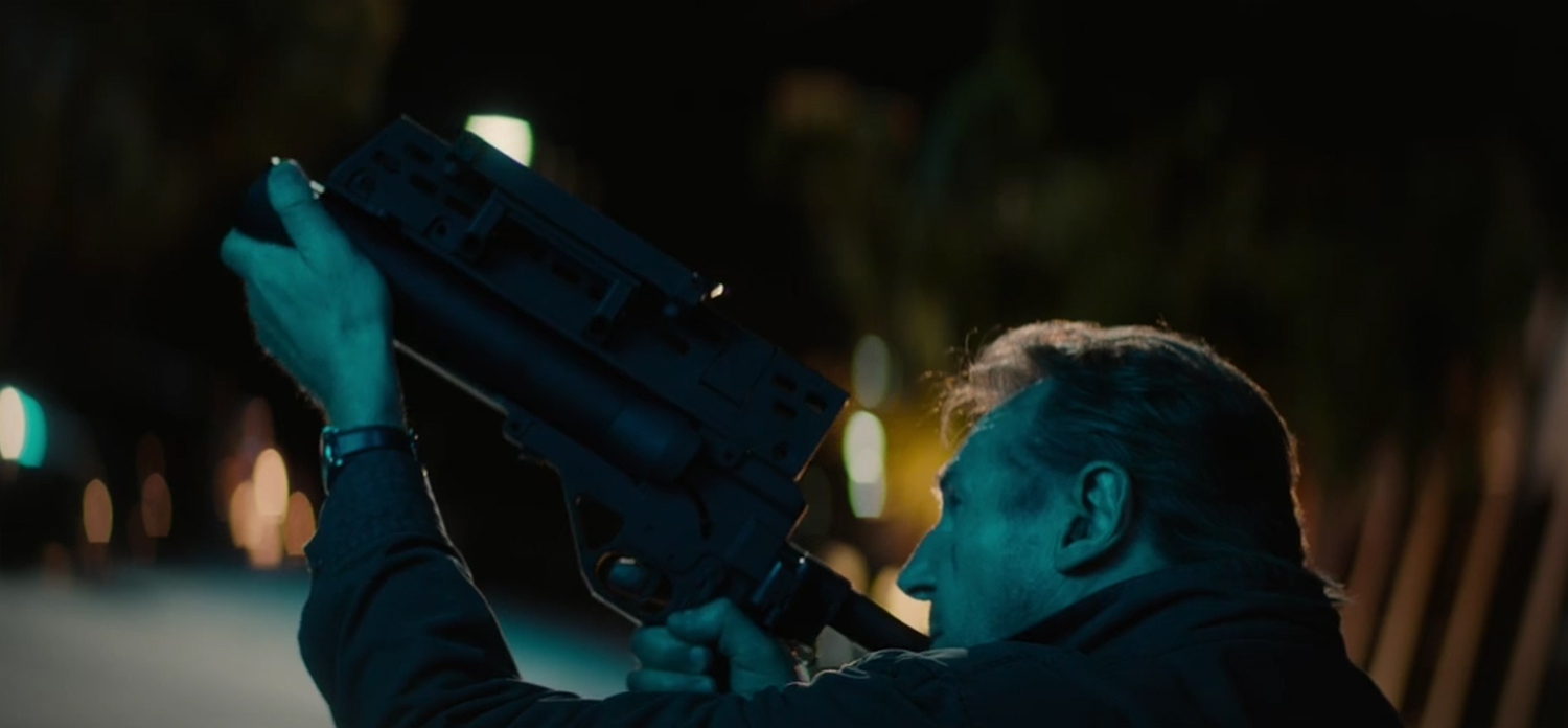 Liam Neeson fires a grenade launcher.
