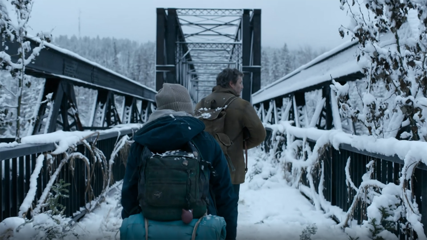 Joel and Ellie walk over a snowy bridge.