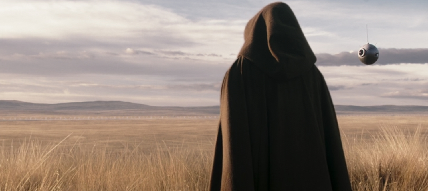 A hooded figure looking over a grass field.