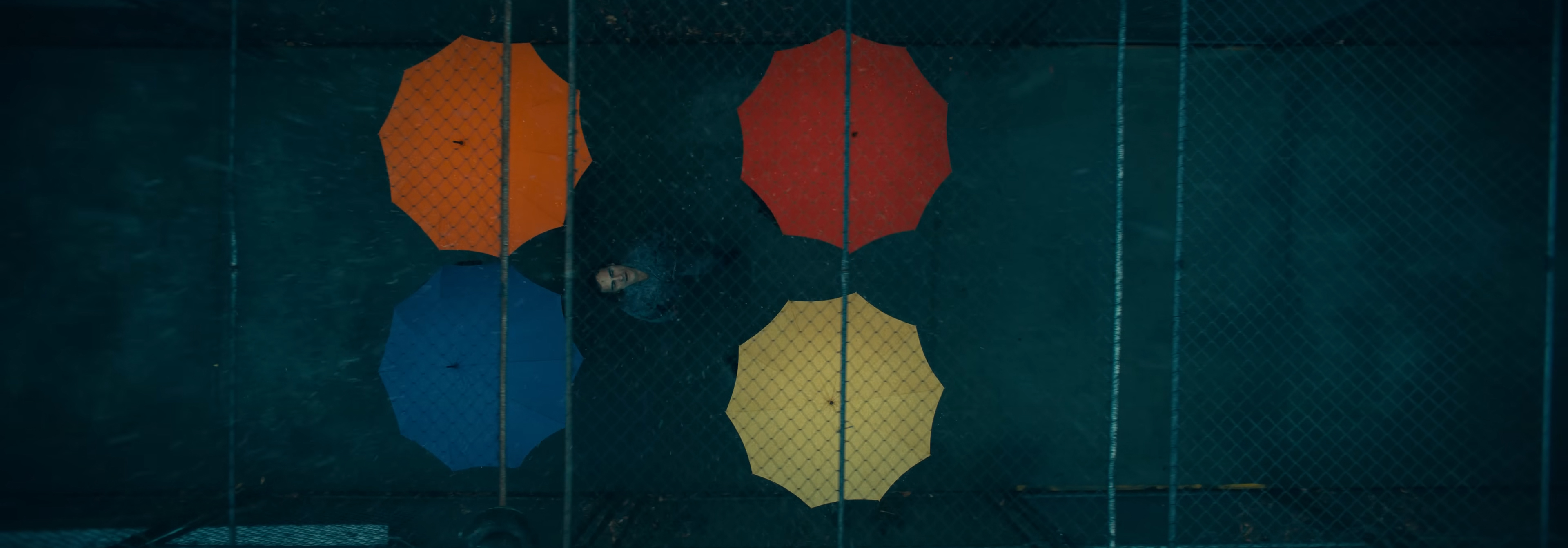 The Joker walks under rain surrounded by colourful umbrellas.