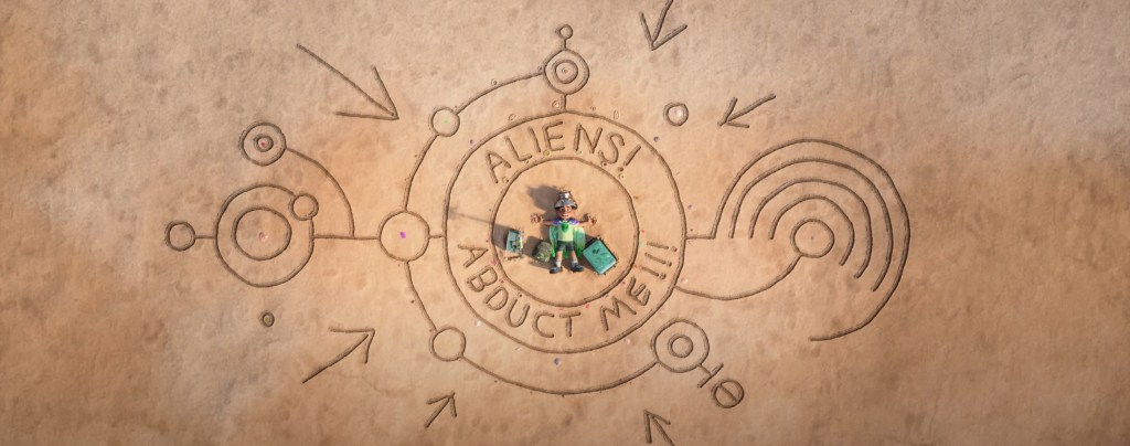 Elio sitting in an 'abduct me' sign written in the sand.