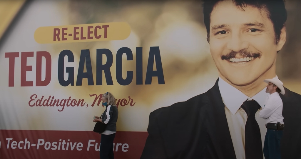 Joaquin Phoenix stands in front of a poster of Pedro Pascal.