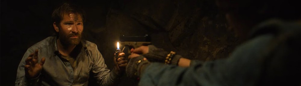 A woman holds a man up with a gun.