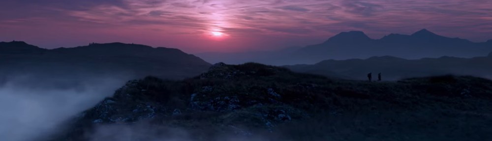 Two men walk on a hill crest as the sun sets behind them causing a purple sky.