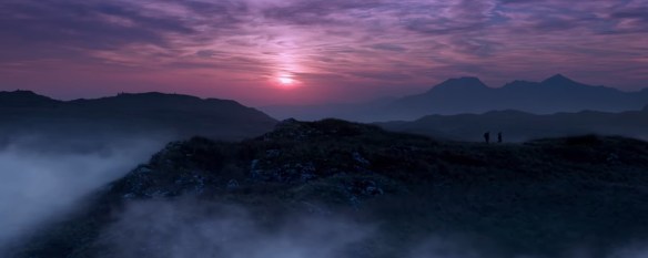 Two men walk on a hill crest as the sun sets behind them causing a purple sky.