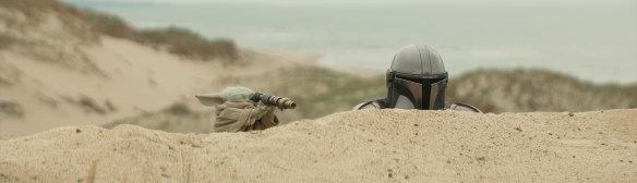 Grogu and The Mandalorian look over a sand dune.