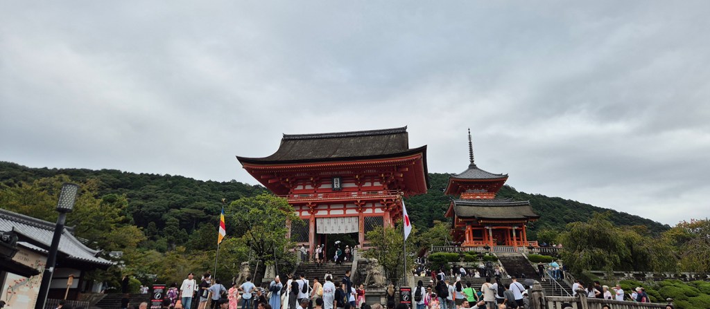 A scenic view of two traditional Japanese temple structures surrounded by lush greenery, with many visitors exploring the area under a cloudy sky.