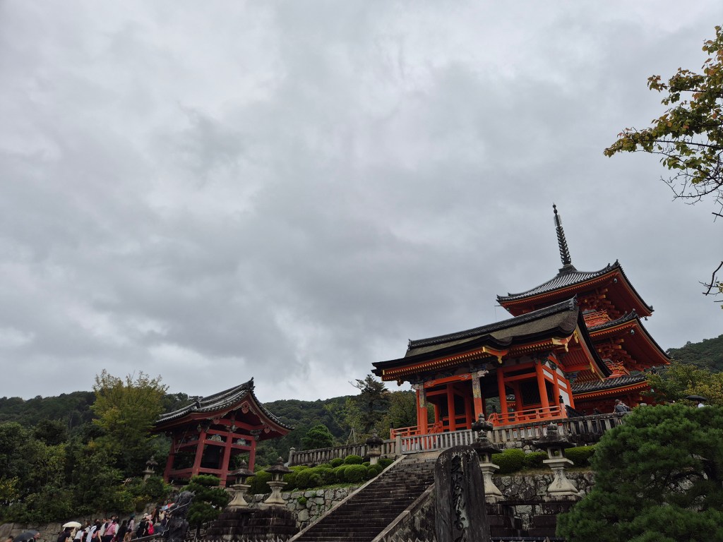 A traditional Japanese temple structure with vibrant red and black colors, set against a cloudy sky. Visitors can be seen ascending steps, surrounded by greenery and stone pathways.