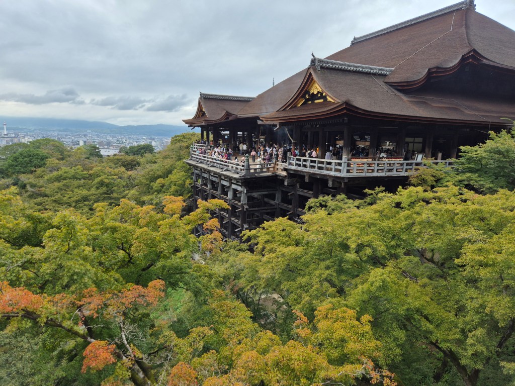 A scenic view of Kiyomizu-dera temple, featuring its wooden balcony supported by stilts, surrounded by lush greenery and trees showing autumn colors. People are gathered on the balcony, looking out over the landscape.