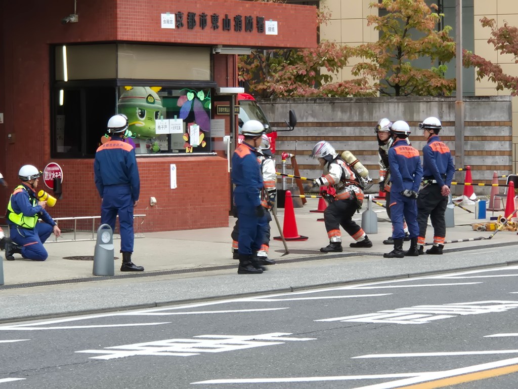 A group of firefighters and emergency responders in uniforms are gathered outside a fire station, some wearing helmets and safety gear, while others are using equipment and communicating. There are traffic cones and a vehicle visible in the background.