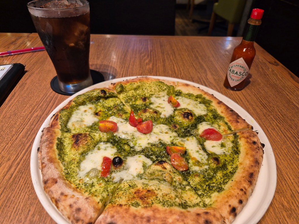 A plate of pesto pizza topped with mozzarella cheese and cherry tomatoes, accompanied by a glass of iced cola and a bottle of Tabasco hot sauce on a wooden table.