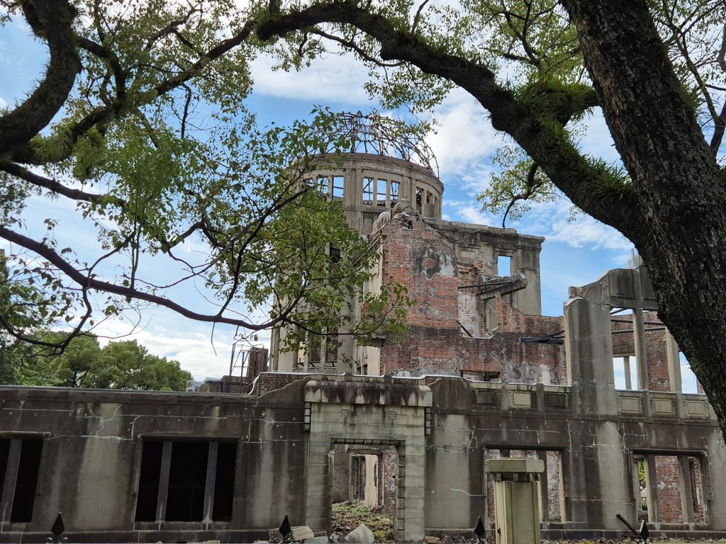 Ruins of a historic building with exposed brick and a dome structure, surrounded by trees and under a blue sky.