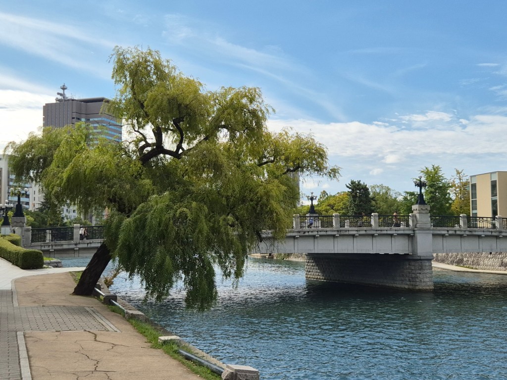 A picturesque scene featuring a bridge over a river, with a weeping willow tree leaning towards the water. Modern buildings and greenery can be seen in the background under a clear blue sky.