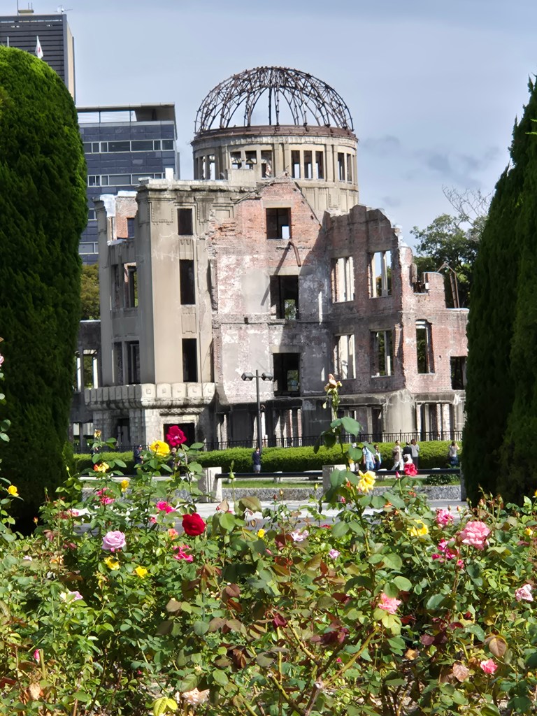 The ruins of the Hiroshima Peace Memorial, with flowering bushes in the foreground, showcasing a mix of vibrant roses against the backdrop of the historical site.