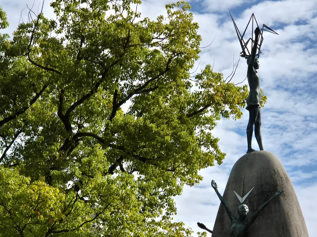 A bronze statue of a figure holding up geometric shapes, perched on a large stone, surrounded by green trees and a partly cloudy sky.