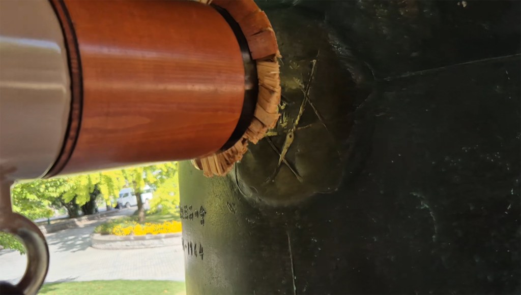 Close-up view of a bronze bell with a wooden mallet resting against it, showing intricate details of the bell's surface and markings.