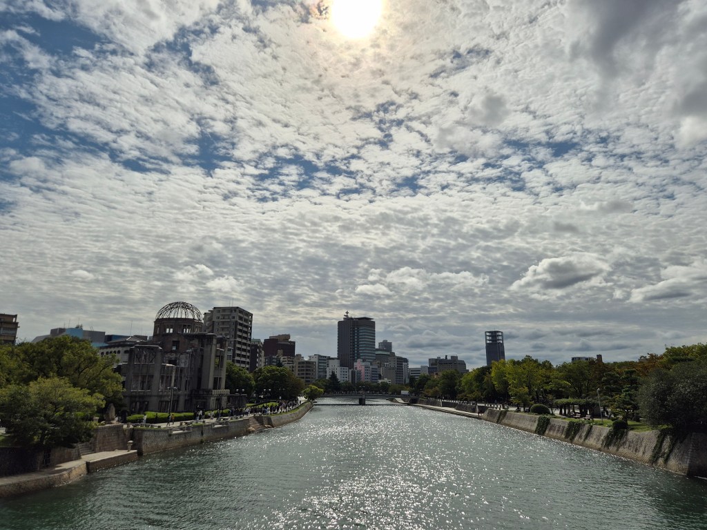 A scenic view of a river running through a city, with a partially cloudy sky and the sun shining in the background. The image features greenery along the riverbanks, modern buildings in the distance, and the ruins of a historical structure visible on the left.