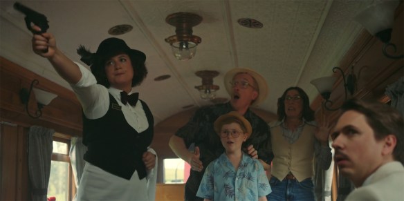 Rose Matafeo holding a gun as a family look on in surprise.