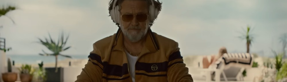 Russell Crowe sitting down in an yellow jumpsuit listing to headphones.