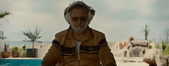 Russell Crowe sitting down in an yellow jumpsuit listing to headphones. 